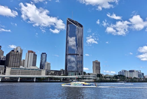 Skyscraper in Brisbane skyline reflecting in river under blue sky with clouds.